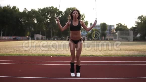 动作缓慢肌肉发达的女人穿着黑色比基尼跳跃在户外体育场使用跳绳正面观点视频的预览图