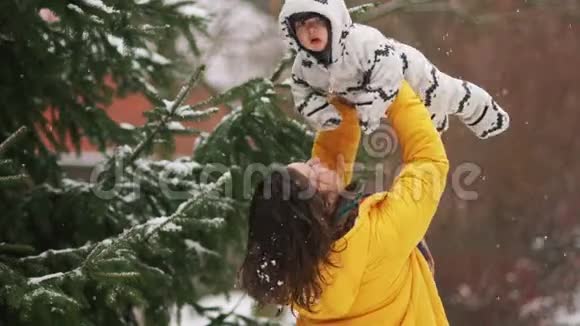 年轻的母亲亲吻她的孩子一个女人抱着她的小儿子在雪下的冬季公园里散步视频的预览图