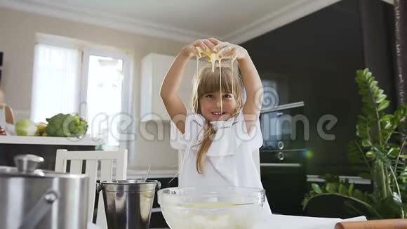 可爱的小女孩一头长发穿着白色连衣裙在玻璃碗里揉着面团在现代厨房里看着相机视频的预览图