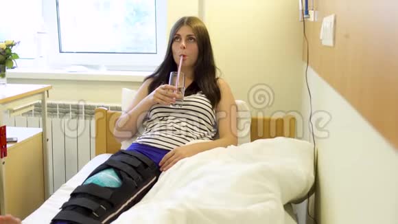一位腿部受伤的年轻病人在医院房间里饮用水和使用电话视频的预览图