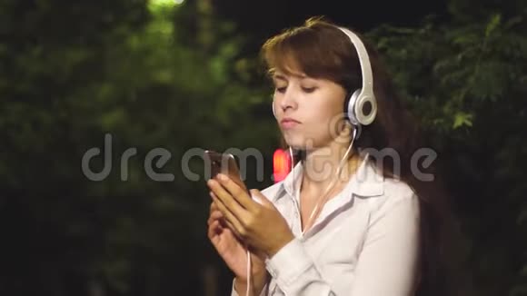 戴着耳机和智能手机的年轻女孩在一个城市公园里听音乐跳舞和微笑特写视频的预览图