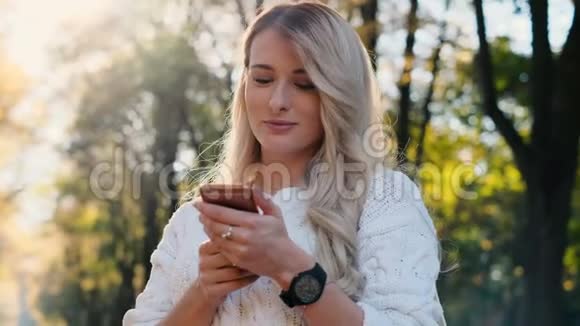 可爱的女孩穿着白色毛衣在她的智能手机户外写信息女人用数码小玩意滚动视频的预览图