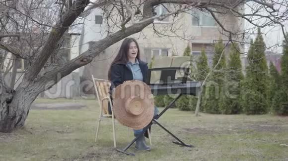 在后院的树下坐着成功的天才美丽的黑发女孩在玩合成和唱歌视频的预览图