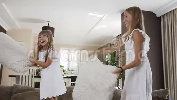 两个穿着白色连衣裙的妹妹跳在沙发上玩得很开心在家里的客厅里用柔软的枕头打架两个可爱的视频的预览图