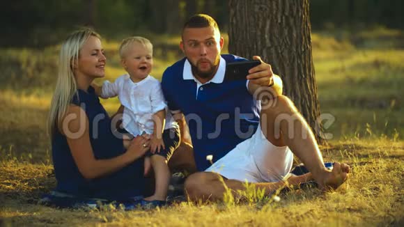 幸福的一家人坐在草地上在公园日落时和一个婴儿一起自拍爸爸和妈妈拍照视频的预览图