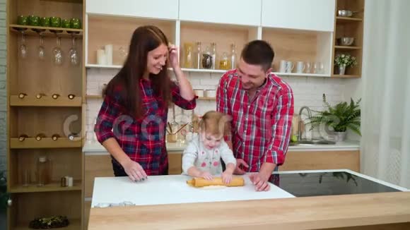 年轻的父母和蹒跚学步的女儿一起玩一起在厨房里擀面团视频的预览图