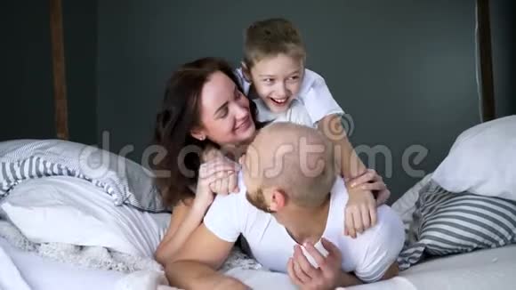 爸爸妈妈和儿子穿着相同的白色T恤衫在室内的床上特写快乐的时刻玩得很开心视频的预览图