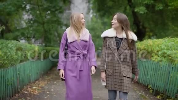 两个快乐的女朋友穿着暖和的外套在秋日漫步在公园的小巷里聊天视频的预览图