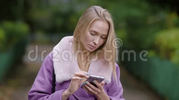 成年金发女商人独自站在公园里白天在智能手机上输入信息视频的预览图