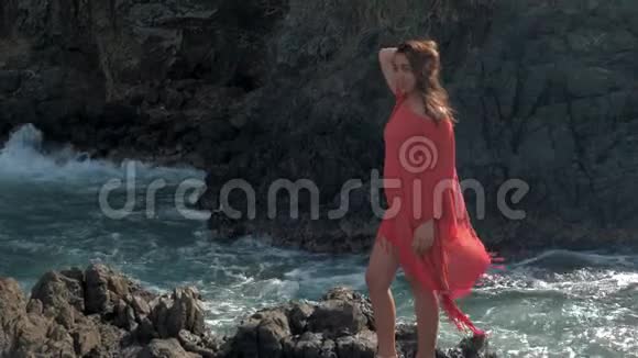 年轻的女孩穿着红色的衣服站在暴风雨的附近海浪拍打着岩石年轻的女孩望着大海海浪拍打着视频的预览图