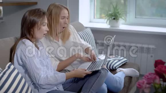 两个女孩坐在家里的沙发上在网上购物在笔记本上讨论和拿着笔记本电脑视频的预览图