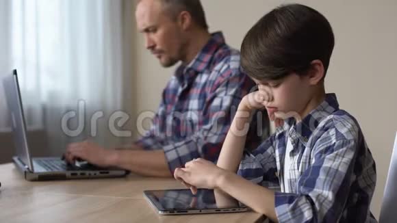 父亲缺乏注意力孩子在平板电脑上玩游戏而父亲使用笔记本电脑视频的预览图