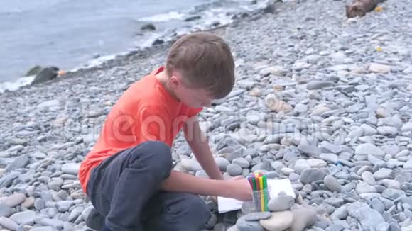 男孩坐在海边的石滩上画着记号笔视频的预览图