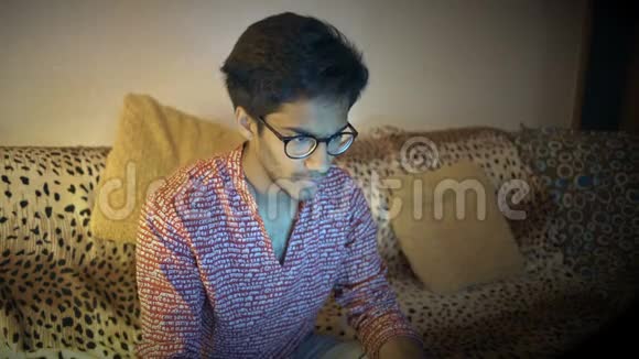 戴眼镜的印度男人坐在家里的沙发上为笔记本电脑工作视频的预览图