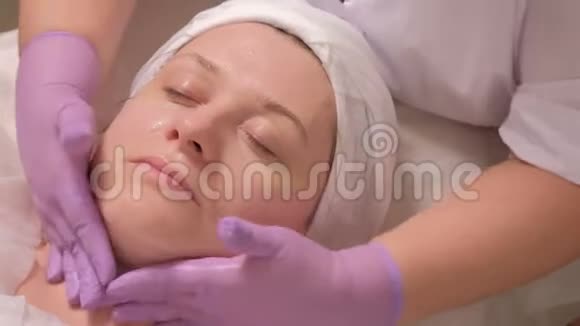 一位戴手套的专业美容师的双手正在美容院客户的女性眼睛周围涂抹眼霜关闭视频的预览图