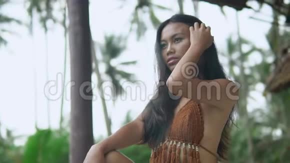 棕色上衣的超级年轻黑发亚洲模特坐在丛林小屋附近看着摄像机视频的预览图