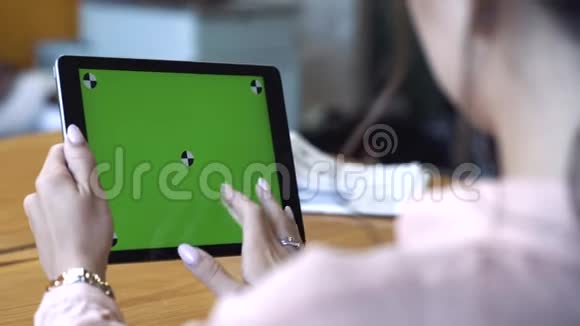 从一位身穿粉红色衬衫的年轻女子的肩膀上看她坐在桌子旁在Chroma键的平板上滑动着视频的预览图