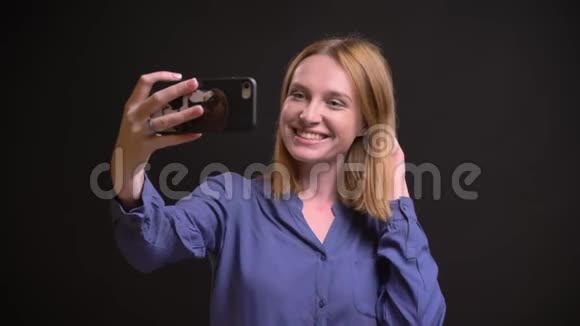 白种人金发女人穿着衬衫在黑色背景下使用智能手机制作自拍照片的肖像视频的预览图