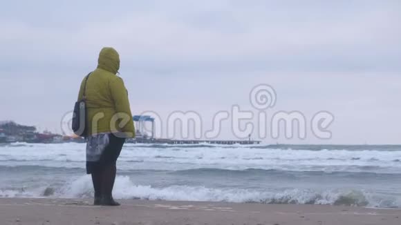 在日落时沙滩上的暴风雨中穿着绿色夹克的胖女人看着海浪后景视频的预览图