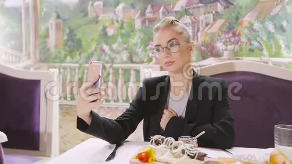 一个年轻女孩坐在餐厅的桌子旁在智能手机上自拍视频的预览图