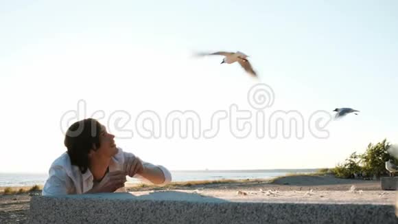 一个穿着白色衬衫的欧洲年轻人在日落时在海边用面包喂海鸥帅哥躲在后面视频的预览图