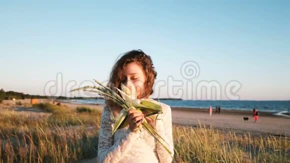 穿着婚纱的年轻欧洲女孩正在嗅着夏天日落时在海边手工制作的一束新娘一个视频的预览图
