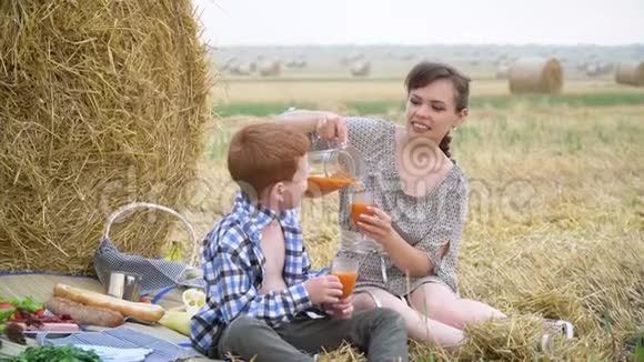 美丽的年轻女子带着红头发的儿子在干草堆中间的野餐中倒灌胡萝卜汁视频的预览图