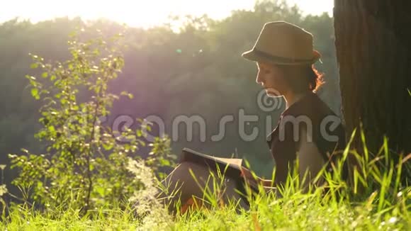 一个年轻的女学生坐在树下的公园里看书户外教育和阅读视频的预览图