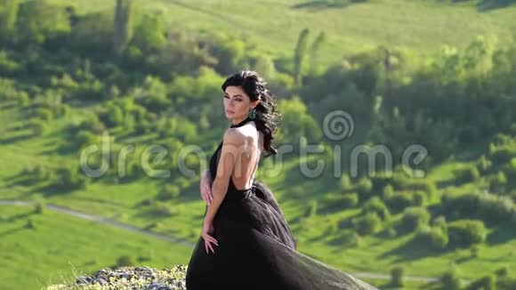一个黑衣碧眼的年轻女子站在悬崖顶上在绿谷的背景下摆姿势视频的预览图