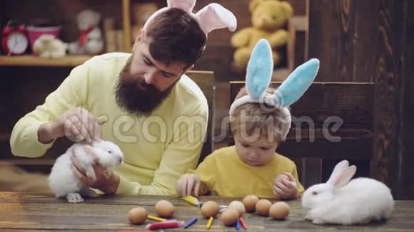 父子俩正在画复活节彩蛋可爱的小男孩戴着兔子耳朵木制背景的复活节彩蛋男人视频的预览图