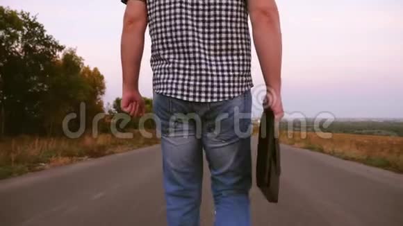 穿格子衬衫的男商人手里拿着一个黑色公文包沿着柏油路走着特写视频的预览图