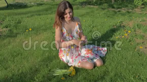 可爱的女人坐在绿色的草坪上编织着一个花圈蒲公英视频的预览图