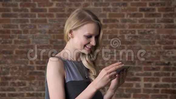 年轻可爱的金发女孩在智能手机上看照片微笑交流理念砖背景视频的预览图