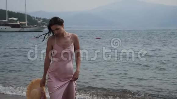 穿着粉红色丝绸裙子和草帽的女人在海滩上散步视频的预览图