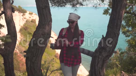一个黑发女孩穿着一件黑色和红色的衬衫戴着头巾戴着眼镜站在树间在电话里拍了张照片视频的预览图