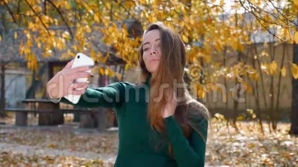 深褐色头发的女人在秋天的黄树背景下在智能手机上自拍视频的预览图