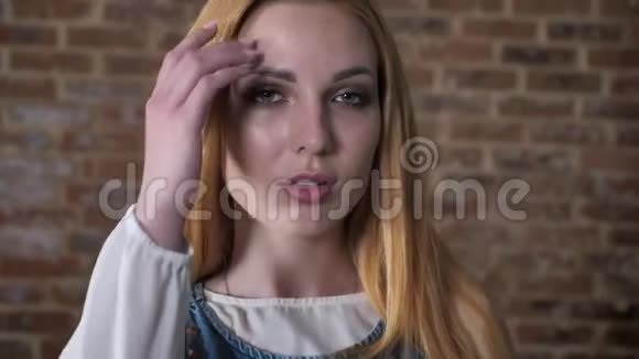 年轻自信的金发女孩在镜头前观看触摸头发调情的概念砖块背景视频的预览图