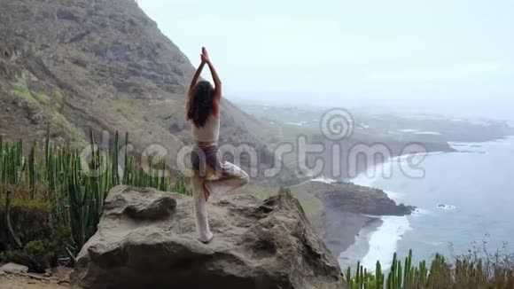 年轻女子在山上做瑜伽在一个俯瞰大海的小岛上站在一条腿上举起双手视频的预览图