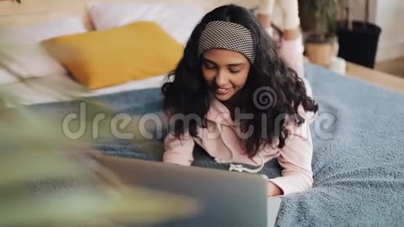 快乐的非裔美国女人躺在床上用笔记本电脑浏览网页穿着粉色睡衣的女孩微笑着视频的预览图