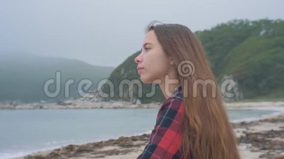 阴天穿着方格衬衫的放松的欧洲女孩坐在沙滩上视频的预览图