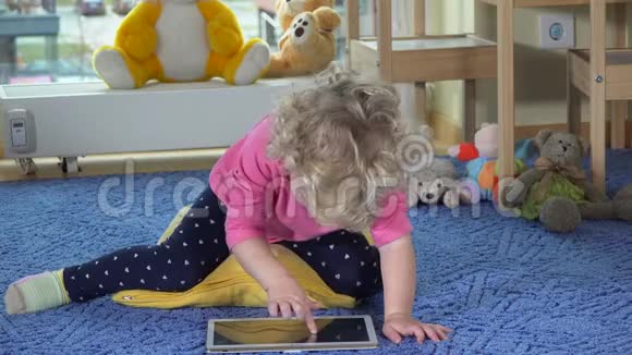 可爱的蹒跚学步的女孩使用平板电脑坐在蓝色地毯上快乐的孩子拍手视频的预览图