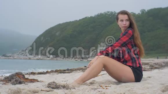 阴天穿着方格衬衫的放松的欧洲女孩坐在沙滩上视频的预览图