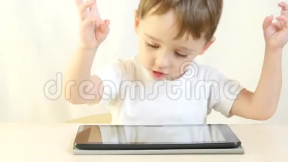 快乐的小男孩坐在桌子旁玩平板电脑一个孩子在玩电子游戏挥舞着手臂因特网和视频的预览图