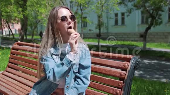 年轻女孩坐在长凳上手上拿着冰淇淋穿着白色T恤牛仔布牛仔裤和太阳镜视频的预览图