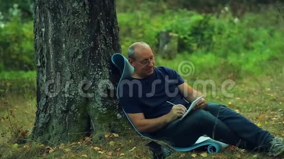 一个戴眼镜的微笑男人坐在公园的一棵树下用铅笔在笔记本上写字视频的预览图