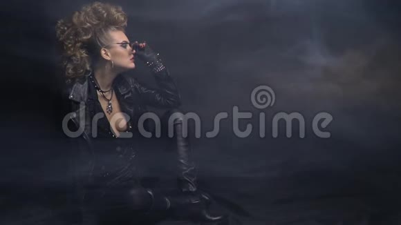 身穿皮夹克的摇滚女人戴着太阳镜烟雾和黑色背景在地板上摆姿势视频的预览图