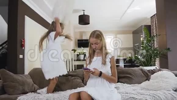 一个穿着白色连衣裙的小女孩跳来跳去玩着一个柔软的枕头姐姐坐在沙发上视频的预览图