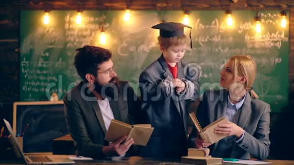 一个穿着夹克和毕业帽的男孩和两个在学校班级背景下拿着书的老师交流视频的预览图
