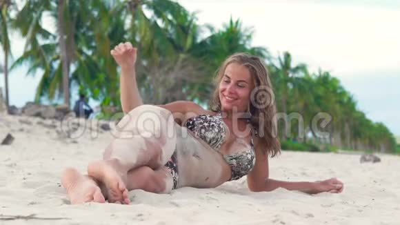 美丽的女人穿着比基尼躺在沙滩上在你的身体上倒沙女人把沙子撒在晒黑的身上视频的预览图