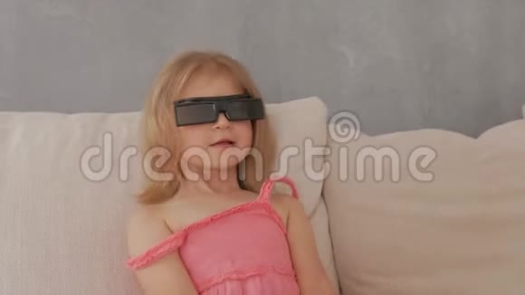 一个穿着粉红色裙子的小女孩坐在沙发上用3D眼镜专注而有感情地看电视视频的预览图